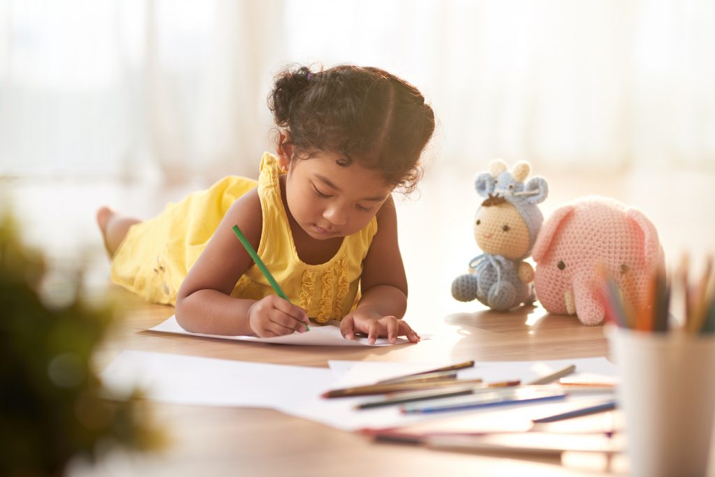 Concentrated toddler in yellow dress coloring picture with pencils while lying on floor, blurred background