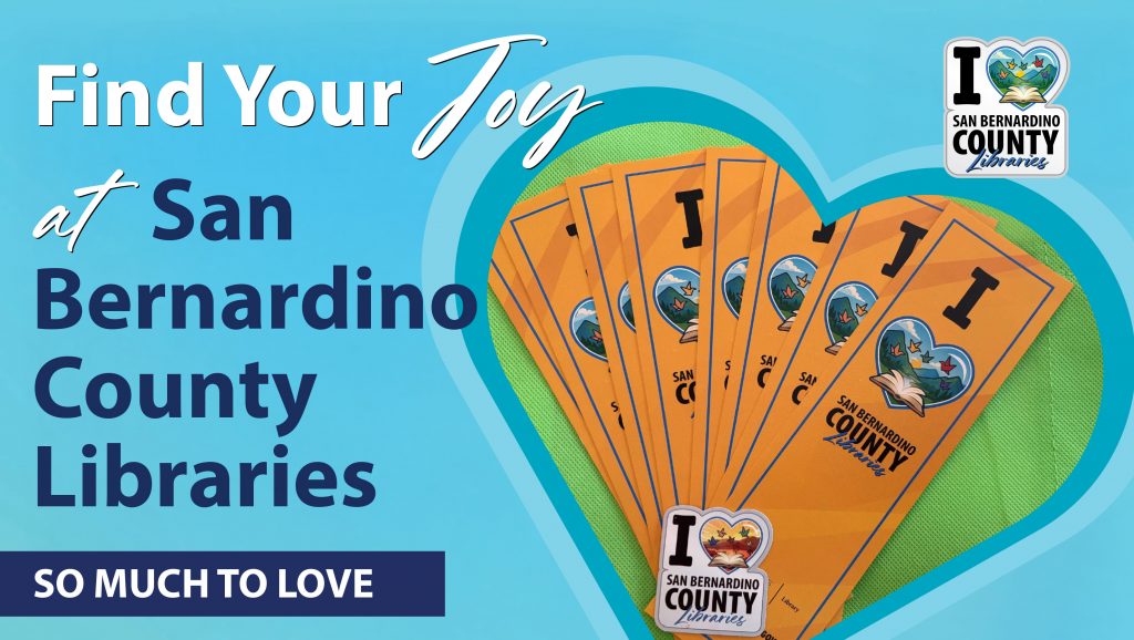 A blue promotional banner on a computer screen reads “Find Your Joy at San Bernardino County Libraries” with the phrase “So Much to Love.” The design includes the San Bernardino County Libraries logo and a heart-shaped graphic filled with a fan of orange bookmarks featuring the library logo and “I ❤️ San Bernardino County Libraries.”